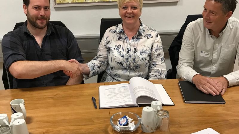 to menn og en dame smiler og håndhilser etter signert kontrakt på skrivebordet foran seg